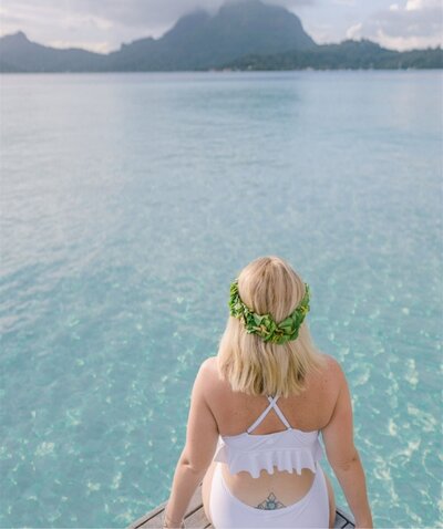 Woman in a white bikini sits on the edge of an overwater bungalow and gazes out at the blue water