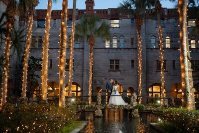Aerial view of a smiling bride in a white wedding dress with flowing veil sitting on brick pavement next to her groom in naval dress uniform with captain's hat, surrounded by lush greenery including ferns and flowering plants at the White Room's Champagne Courtyard