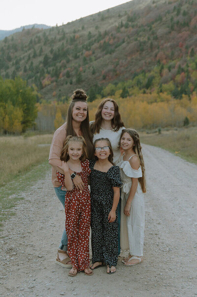 A mother poses with her 4 daughters, all smiling at the camera. They standing on a dirt road with lots of foliage on a hill far behind them.