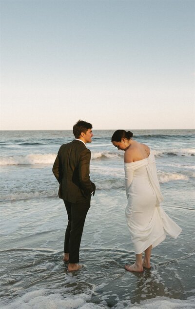 A moment captured by a Wilmington wedding photographer of newlyweds walking barefoot in the low tide waters on the beach at sunset