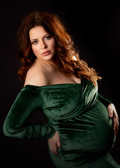 A mother to be stands in a studio gazing over her shoulder in a velvet green maternity gown