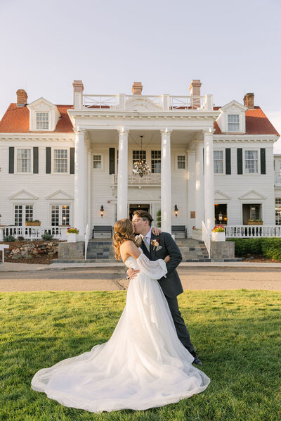Bride and groom sharing a kiss in front of The Manor House, a classic Colorado wedding venue with white columns and historic charm.