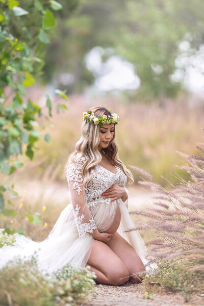 Pregnant woman kneeling in a field in a white maternity dress with a lace top and tulle bottom wearing a floral crown and holding her belly