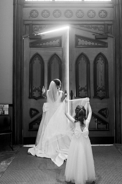a girl holding a bride's train while she walks in a cathedral