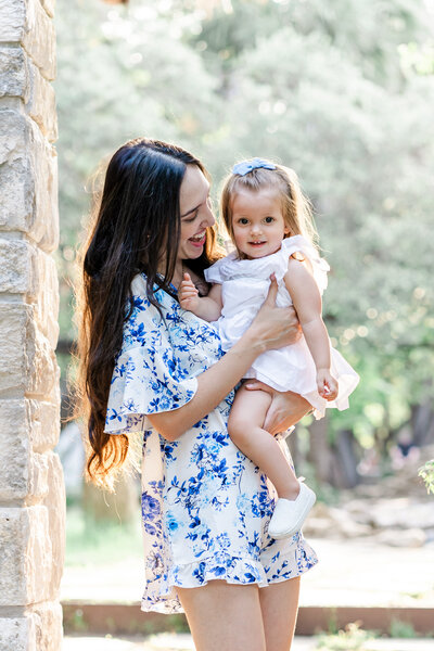 A mother stands under an arch at Mayfield Park in Austin, TX. She holds her daughter in her arm, dress in a white dress with a blue bow in her hair. The little girl smiles at the camera while the mother looks at her smiling.