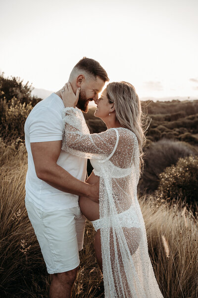 A couple sits closely on a sandy beach at sunset, gazing at each other lovingly, captured beautifully by an Illawarra maternity photographer.