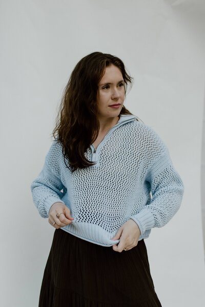 A woman in front of white backdrop fixing her blue sweater looking to the right. 