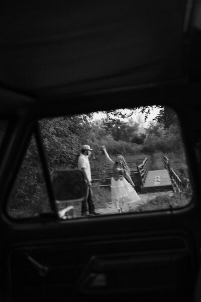 View of engaged couple dancing in field through the window of Ford truck