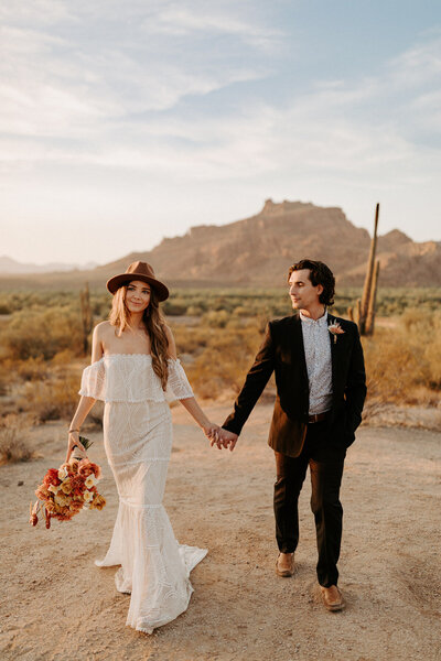 A bride and groom in wedding attire walk towards the camera. The Phoenix desert is seen in the background with cacti and a tall mountain far in the distance. The sun is setting to their left. The bride wears a lace off-shoulder dress that flows down to her feet. She wears a brown hat and is holding an orange bridal bouquet. Her left hand is  being held by her groom. He wears a black suit with the suit jacket unbuttoned.  A white floral button-up can be seen underneath. He has a boutonniere on. He is happily looking at his bride.