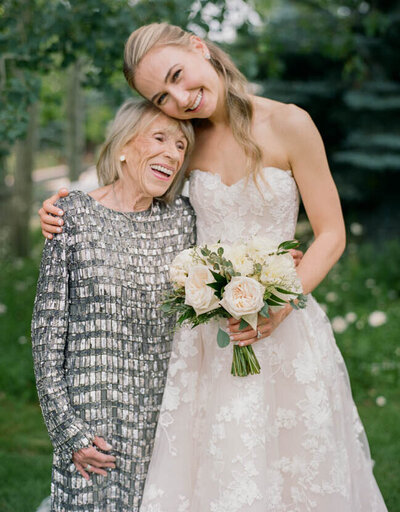 A bride in a white floral gown smiles while holding a bouquet and standing beside an elderly woman in a silver dress. They are outdoors with greenery in the background.