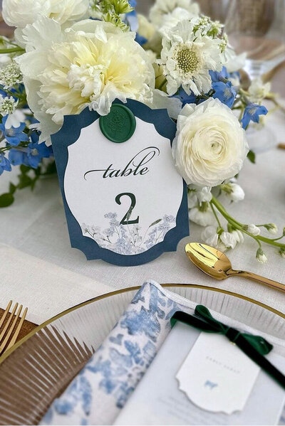 French blue velvet wedding table number with a forest green wax seal nestled within a blue and white floral centerpiece, showcasing sophisticated wedding invitation design.