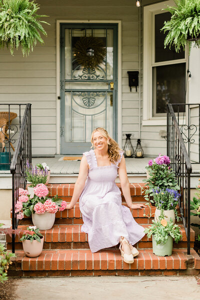 A female DIYer wearing a purple dress sits on the front porch of her victorian home surrounded by potted plants in bloom.