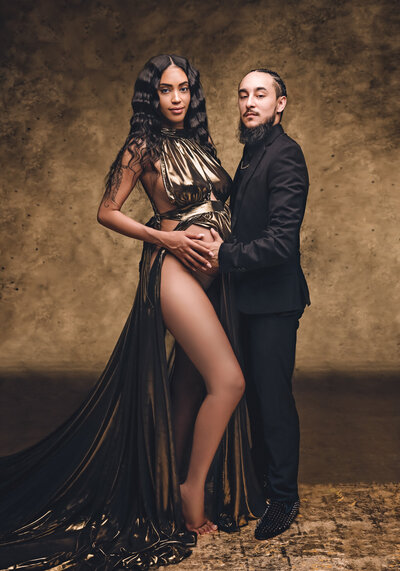 A couple poses against a warm, textured brown backdrop. The woman, wearing a metallic gold gown with a flowing train and high slit, stands close to the man clad in a black suit. He embraces her pregnant belly, symbolizing love and expectancy. Her long, wavy hair contrasts elegantly with the rich gold and earthy tones of the image.  