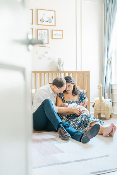 A couple sits on the floor of their baby's nursery. The father wraps his arm around the mother while she holds their newborn. They both look down at their baby. 