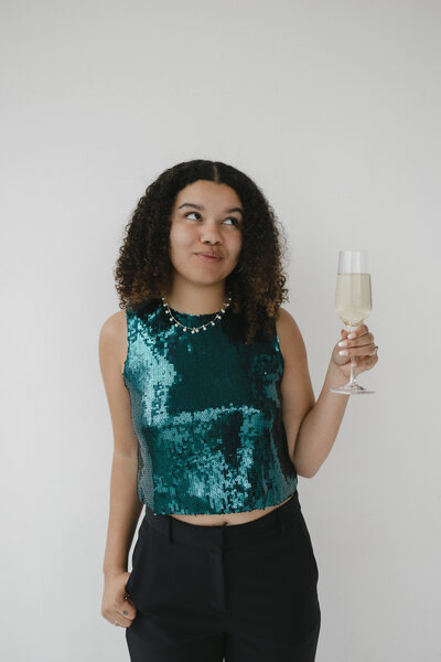 Photo of Catryce holding champagne in a teal shirt with a neutral background