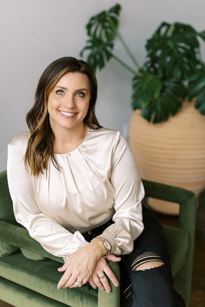 A woman with long brown hair, wearing a cream blouse and ripped black jeans, sits on a green chair. She smiles at the camera, hands resting on her lap. A large potted plant with broad leaves is in the background.