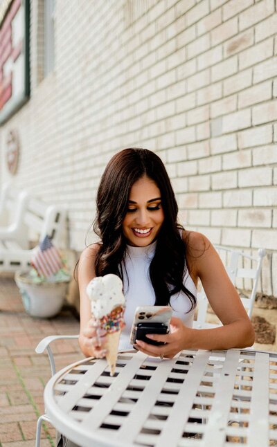 fashion show nail tech sitting and holding ice cream on one hand and phone on the other hand while smiling