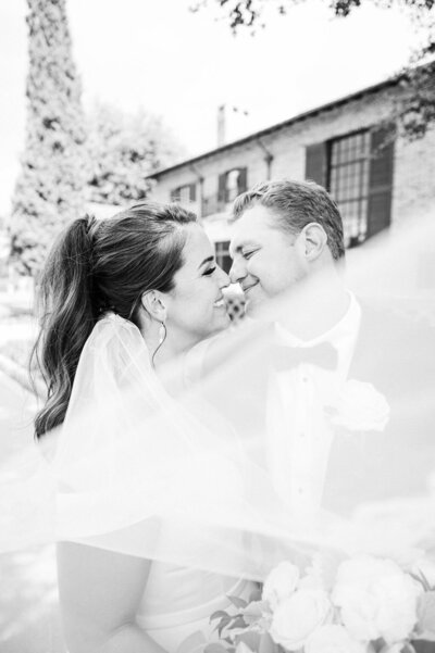 A bride and groom share an intimate moment, with the bride’s veil flowing in the breeze, captured in an artistic black-and-white shot.