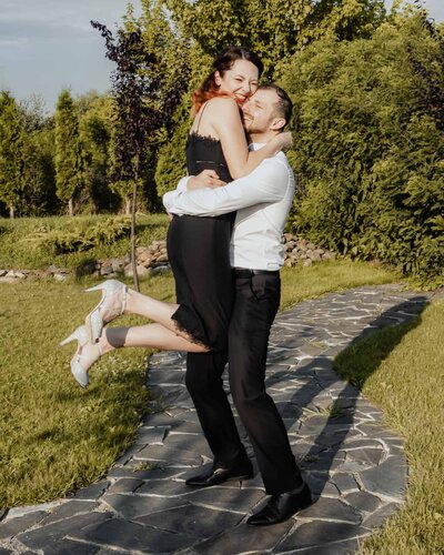 A smiling couple embracing outdoors on a sunny day, with the woman joyfully lifted off the ground as they share a fun and romantic moment, captured by a UK wedding photographer and videographer.