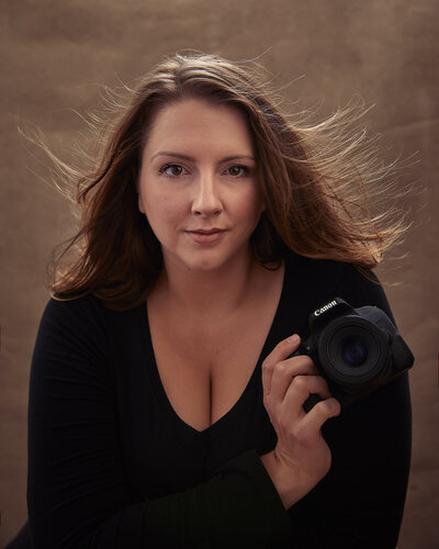 Self-portrait of Norwich Portrait photographer Claire Howes. She is holding a Canon camera and looking at the camera with her hair flowing behind her. She is posed on a brown canvas background in her studio in Norwich.