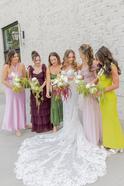 Bride gathers with her bridal party, in assorted colored dresses and beautiful wedding bouquets, all looking at the bride and smiling, having a good time in Raleigh, NC at the Maxwell.