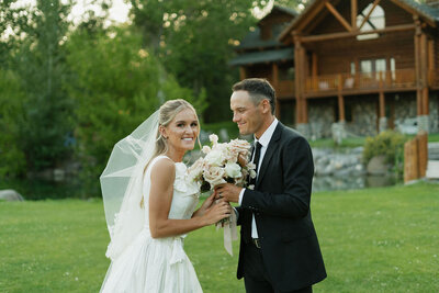 A bride and groom hold the bride's wedding bouquet together. She is looking at the camera smiling and he is looking at her., both facing each other. Behind them is a large wooden cabin and trees.