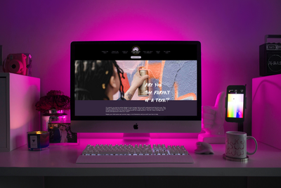 This image shows the Teen Therapy page of the Inspired Mental Wellness displayed on an iMac, surrounded by colorful desk clutter. The purple of the website is highlighted by a purple light emanating from behind the computer.
