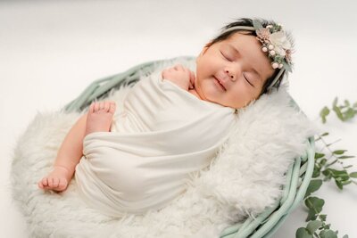 Newborn baby wrapped in a soft white swaddle, peacefully sleeping on a fluffy white blanket in a round basket with a delicate floral headband and greenery accents.