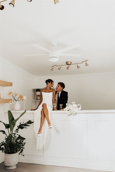 Bride in a modern white dress sitting on a sleek white bar, sharing a joyful moment with the groom at Uliana Estate in Florida