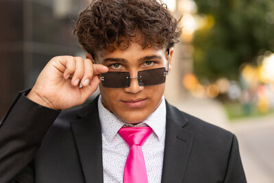 A senior boy poses confidently in a sleek black suit with a hot pink tie, tilting his sunglasses for a bold, fashion-forward look. The blurred city lights in the background add an urban, modern touch to this striking senior portrait.
