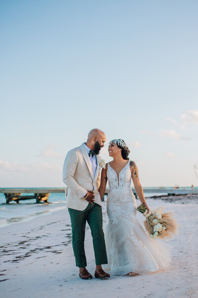 A bride and groom walk hand in hand along the beach. The groom wears a white blazer and green pants, while the bride wears a beaded gown and a jeweled headpiece. heir smiles and relaxed energy capture the romantic, carefree spirit of a destination wedding expertly planned for effortless celebration.