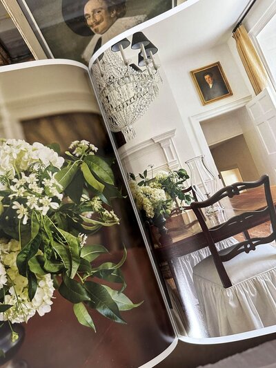 A top-down view of an open book displaying a vase filled with white flowers and greenery on one page, and a beautifully set dining table in a historic home on the other.