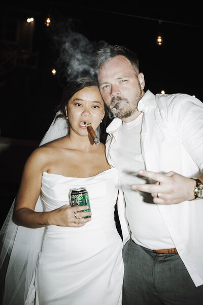 A bride in a strapless white gown and a man in a white shirt pose together at night, both smoking cigars, with the bride holding a can of soda and smiling confidently.