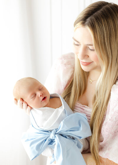 Young mom wearing a pink dress from CCP's client closet holding her newborn baby boy wrapped in a white and blue bow swaddle. She is looking at him with eyes full of love
