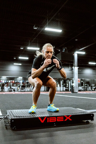 Woman squats on a piece of machinery in a gym setting.