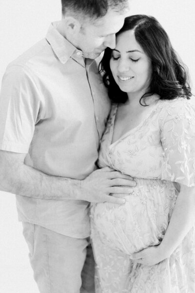 Black and white photo of a pregnant mom in a lace gown softly smiling while her husband snuggles in, hand gently on her baby bump by Boston family photographer Fieldstone Studio.