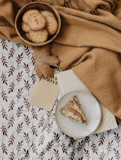 Autumn cozy home scene adorned with a tan waffle blanket, a bed sheet of white with deep plum colored hand painted botanical branches on it, a bowl of maple cookies, a notebook, and a  plated pear tart