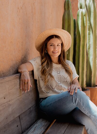 Natalie Bernacchi sitting on a wooden bench in a straw hat and jeans, smiling beside a tall cactus and terracotta wall