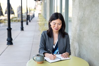 Woman sitting at a cafe table with a coffee cup and writing in a journal.