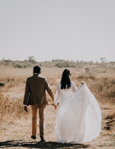 Boho wedding couple holding hands in the desert. Bride and groom in luxury wedding dress and tux. Feels adventurous, trendy, and chic.