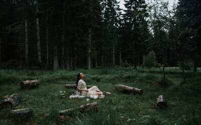 woman is sat on a forest clearing floor as it rains, the trees are huge looming behind her