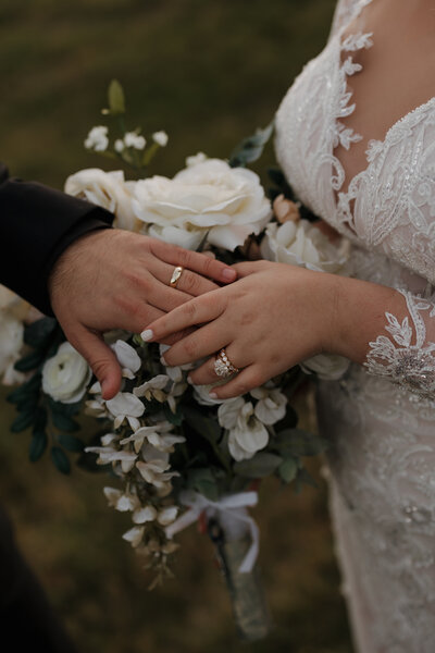 Close-up of a couple's hands adorned with wedding rings. The bride, in a lace dress with intricate patterns, holds a bouquet of white flowers. The groom's hand touches hers gently, as captured by a New England Wedding Photographer against a softly blurred grassy background.
