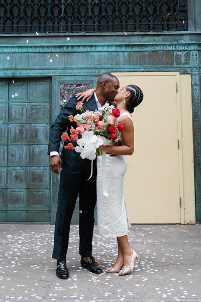 NYC city hall elopement couple kissing outside exit doors with bloom bloom white and red bouquet and before by sarah seven dress