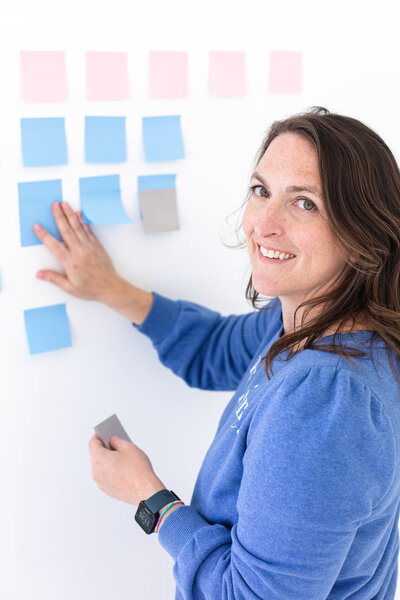 creator of courses for business owners is sticking pink, green and light blue sticky notes with her right hand to the white wall. she is wearing a blue jacket and smiling