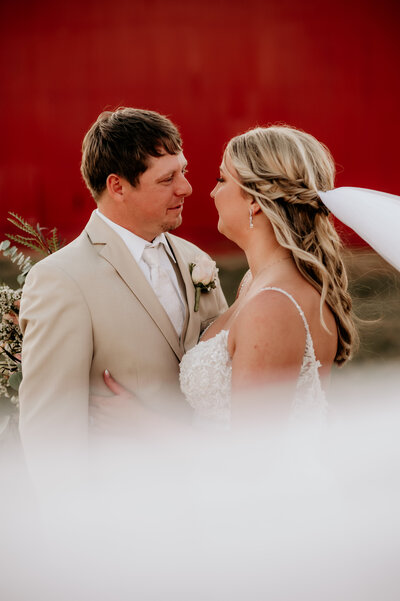 bride and groom standing together in front of a red barn looking into each others eyes romantically as the brides veil blows in the wind