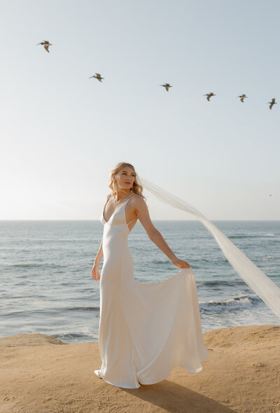 A bride holds out her dress while birds fly behind her at the beach in La Jolla