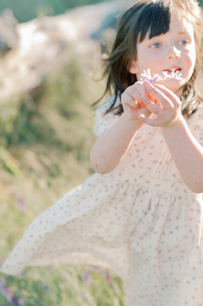 young daughter in a floral dress holds up sweet pea flowers for photograph