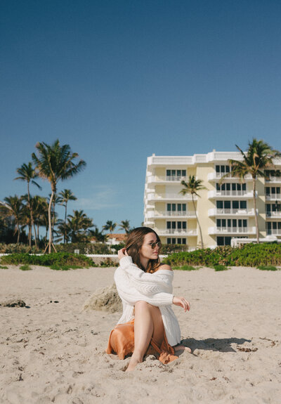 woman sitting on the beach with palm trees and buildings behind her