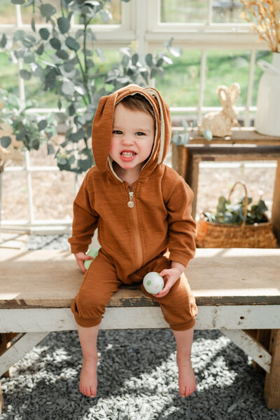 Little boy in a bunny outfit holding an easter egg sits on a bench.