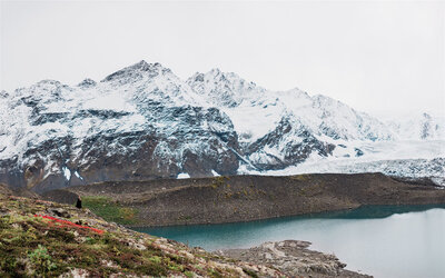 A bride in a black wedding dress is tiny in the frame, standing overlooking a bright blue glacier lake with red and green tundra plants around her, and huge jagged, snow covered peaks that fill the back of the frame.  The spot was access during a luxury glacier elopement package experience in Alaska.